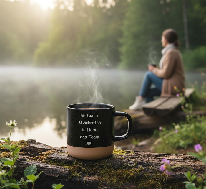 Dampfende schwarze Tasse mit Gravur an einem nebligen See am Morgen